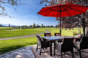 Outdoor patio with a dining table and six wicker chairs under a large red umbrella, overlooking a green golf course and a winding path that inspires movement and new beginnings on a sunny day. Trees and clear sky complete the peaceful scene.