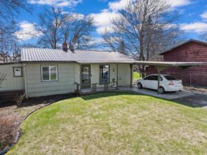 A small, single-story house with light green siding and a metal roof sits next to a carport where a white car is parked. The front yard’s fresh green grass hints at new beginnings, while leafless trees stand still under a blue sky with scattered clouds.