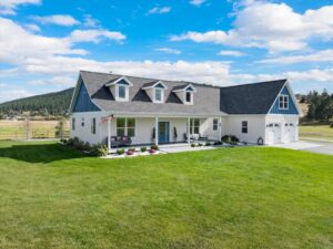 A modern, light-colored house with dormer windows sits on a well-manicured lawn, perfect for relaxing Lake Days. Surrounded by trees and hills under a blue sky with clouds, this rural listing offers peaceful charm and scenic views.