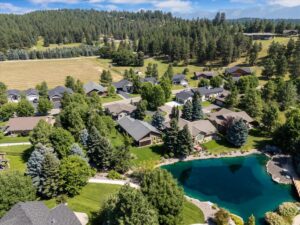 Aerial view of a suburban neighborhood with houses surrounded by tall trees and green lawns, next to a small blue pond, as community lights gently glow against the backdrop of forested hills and fields.