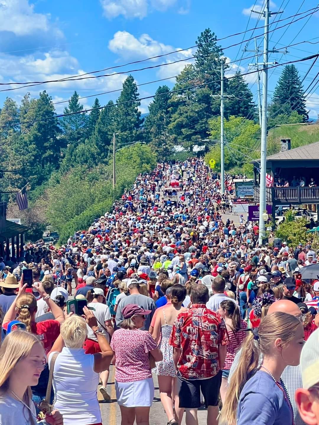 A large crowd of people walks up a sunny, hilly street lined with trees and buildings during the HOT Summer Listing Season, many wearing summer clothes and hats, under a blue sky with scattered clouds. Power lines and signage are visible.