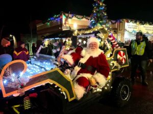 Santa Claus sits in a festively decorated vehicle with Christmas lights, ornaments, and candy canes, smiling at a nighttime holiday parade. Merry Christmas wishes fill the air as people and a tree with lights shine in the background.