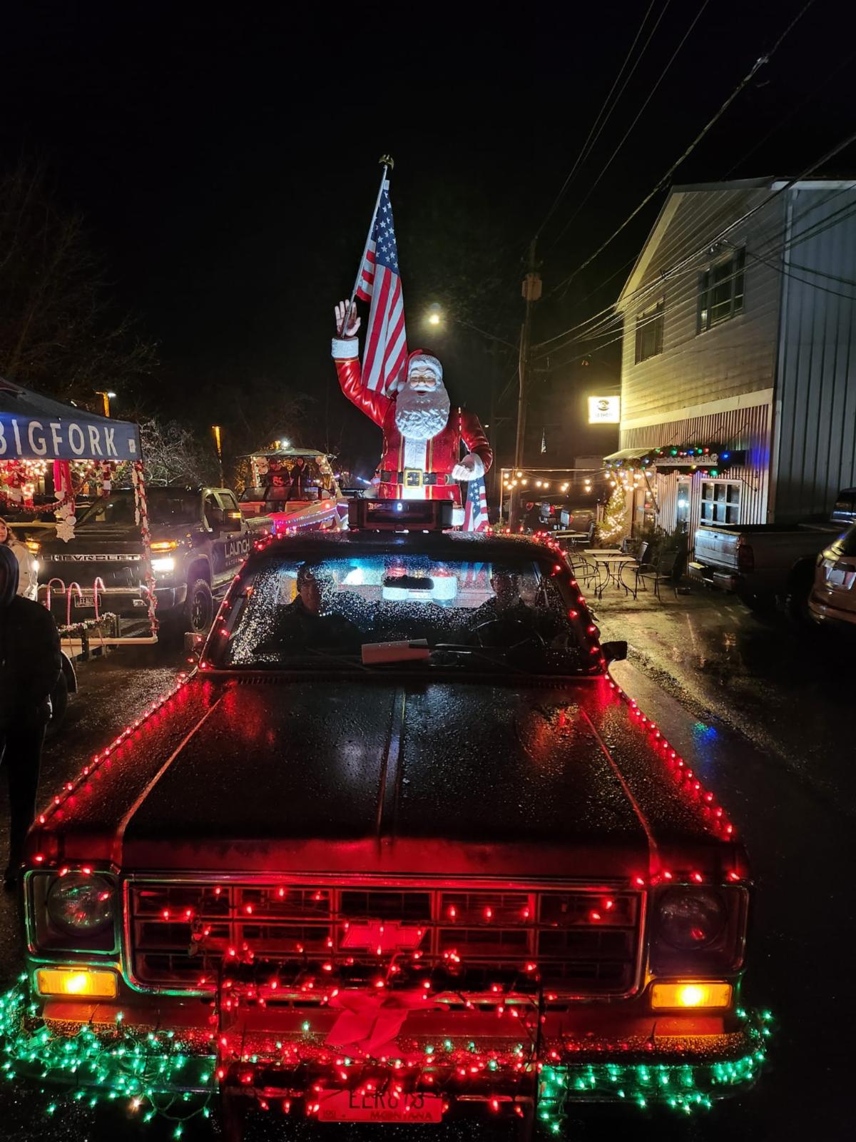 A pickup truck decorated with red and green Christmas lights features a large waving Santa Claus holding an American flag on the roof, spreading Merry Christmas and New Year cheer. The festive night glows with lights and joyful people in the background.