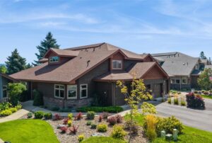 A modern suburban house with brown siding, a gabled roof, and stone accents, surrounded by landscaped gardens and a small tree under a clear blue sky—perfect for your December Real Estate dreams.