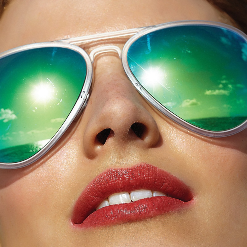 Close-up of a person's face wearing mirrored sunglasses that capture August Reflections of the bright sun, blue sky, clouds, and ocean. The person has red lipstick and fair skin, sunlight highlighting their features in this Season of Action.
