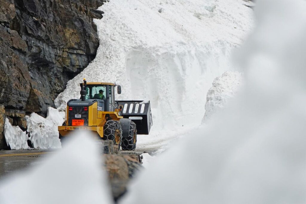 A yellow front loader clears deep snow from a mountain road near rocky cliffs, suggesting ongoing snow removal efforts—an everyday scene before the festivities of Bigfork Days or visits to Blooming Homes begin.
