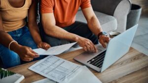 Two people sit on a couch in Flathead Valley, reviewing documents and using a laptop on a coffee table filled with paperwork and a cup of coffee, preparing for the busy Spring Market.