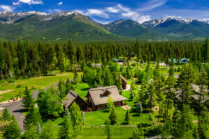 Aerial view of a wooden cabin surrounded by lush green trees, with mountains in the background under a bright blue sky—an inviting scene for anyone exploring Flathead Valley Real Estate.