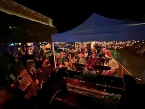 A group of people wearing festive hats sings under a blue canopy on a parade float at night, surrounded by a crowd holding candles or lights, creating a warm, Winter Glow atmosphere.
