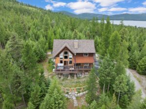 A wooden cabin with a steep roof sits among dense green trees, with mountains and a lake visible in the background under a partly cloudy sky—an ideal retreat for August Reflections and quiet moments.