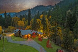 A cozy house surrounded by tall pine trees and autumn foliage sits in a mountain valley at dusk, its warm glow inviting—a perfect retreat for those seeking Flathead Valley real estate under a starry night sky.