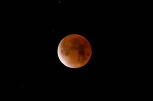 A close-up image of the moon during a lunar eclipse, appearing reddish-orange against a black night sky with a few faint stars—just as breathtaking as a Spring Market evening in Flathead Valley.