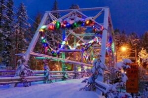 A snowy bridge decorated with colorful holiday lights and garlands, surrounded by snow-covered trees at dusk. A streetlight glows, a “One Car at a Time on Bridge” sign is visible, celebrating a Bright New Year and Merry Christmas.