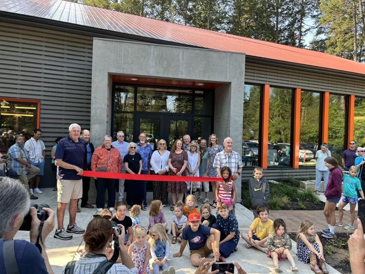 In August, a group of people stands behind a large red ribbon in front of a modern building for a ribbon-cutting ceremony, while children sit on the ground and others take photos. Trees and parked cars are visible as the full month keeps rolling on.