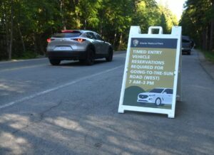 A white sandwich board sign on a road reads: “Timed entry vehicle reservations required for Going-to-the-Sun Road (West) 7 AM–3 PM.” A gray car drives past in a wooded area, highlighting local changes like winter real estate updates.