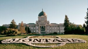 The Montana State Capitol building stands behind a large floral arrangement on the lawn that spells out MONTANA in white flowers, perfect for July Journeys, surrounded by trees under a clear sky.