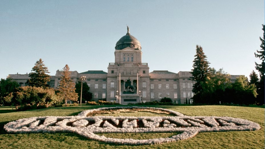 The Montana State Capitol building stands behind a large floral arrangement on the lawn that spells out MONTANA in white flowers, perfect for July Journeys, surrounded by trees under a clear sky.