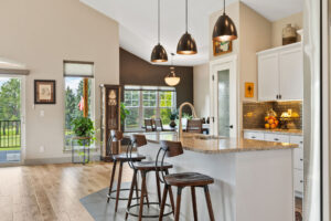 A modern kitchen from our Featured Listings showcases a granite island, three wooden bar stools, pendant lights, white cabinets, stainless steel appliances, and large windows overlooking a green outdoor area.