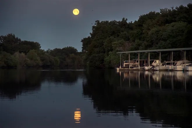 A bright full moon rises over a calm river bordered by trees, its reflection visible in the water. Several boats are docked under a white canopy on the right, creating a scene reminiscent of peaceful Bigfork Days.