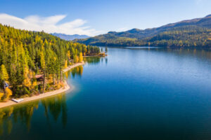 Aerial view of a calm blue lake—Flathead Lake Home—surrounded by dense pine forests and mountains under a sunny sky. The sandy shoreline curves along the forest’s edge, showcasing hints of autumn and the beauty of Flathead Valley Real Estate.