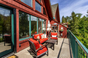 A wooden balcony with red cushioned furniture overlooks a green forest. The deck features a coffee table, umbrella, potted plants, and large windows on a red cabin exterior under a clear blue sky.