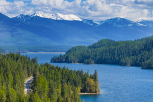 A winding road borders a blue lake surrounded by dense evergreen forests and snow-capped mountains—an idyllic scene showcasing the natural beauty that defines Flathead Valley real estate.