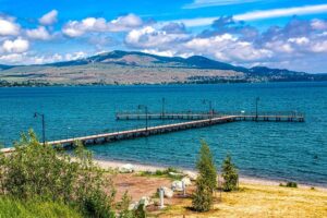 A long wooden pier extends into a blue lake, with distant hills under a partly cloudy sky. Along the shoreline’s greenery and sandy beach, whispers of local folklore linger as dusk brings the gentle glow of community lights.