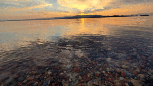 A calm lake at sunset with colorful stones visible beneath the clear water in the foreground and mountains silhouetted against a golden sky, capturing August Reflections in this tranquil Season of Action.