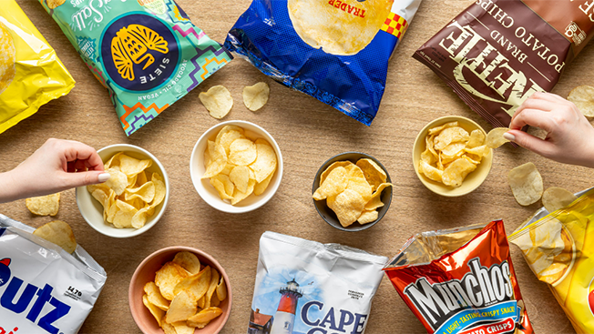 An assortment of potato chip bags and bowls filled with chips are arranged on a wooden table, inviting a sense of community spirit. Two hands reach in to grab chips from different bowls, capturing the warmth of the cozy season.