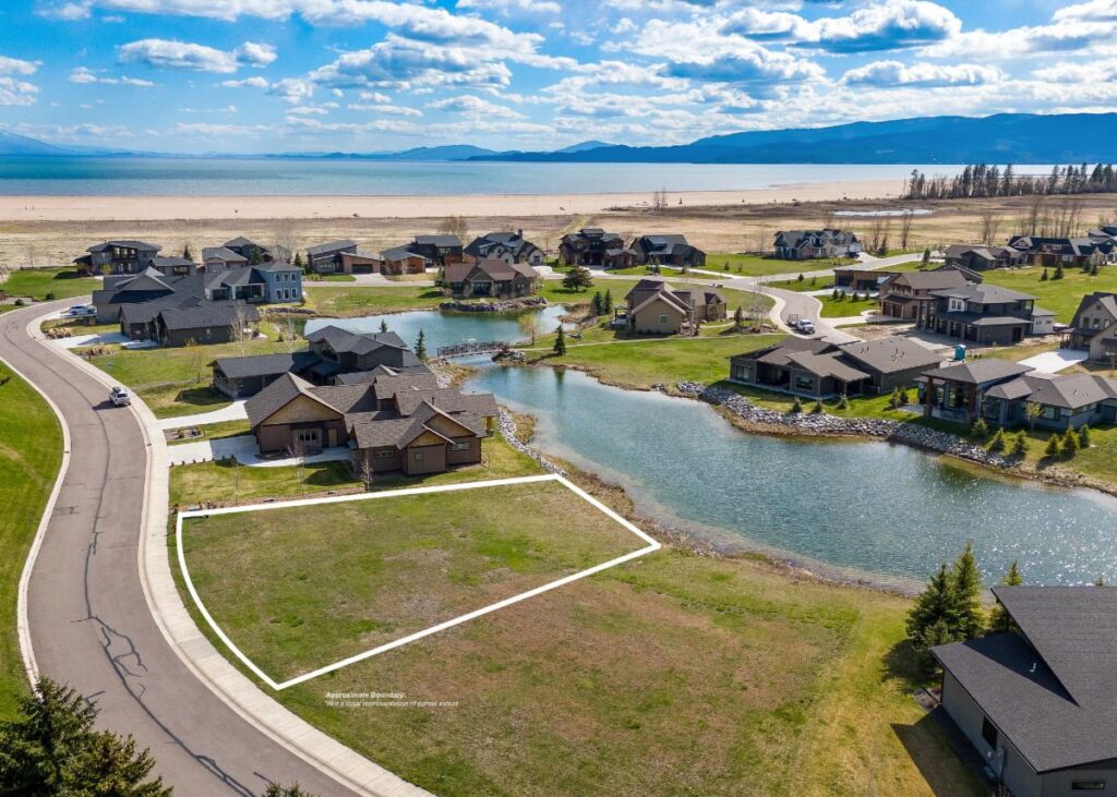 Aerial view of a residential neighborhood with modern houses along a curved street by a pond; one grassy, empty lot outlined in white signals new opportunities, with a beach and large lake beyond under the bright Spring sky.