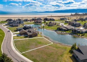 Aerial view of a residential neighborhood with houses by a small pond. A highlighted empty lot with white outline sits in the foreground, next to a curving road, with mountains and a lake in the background on a mild spring day.