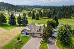 Aerial view of a house with a manicured lawn, driveway, and surrounding trees in Flathead Valley, next to a golf course with lush fairways, sand bunkers, and a small pond—perfect for enjoying the vibrant Spring Market season.