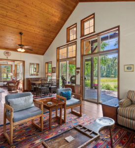 A cozy living and dining area with wood-paneled ceilings, large windows, and glass doors overlooking a green outdoor landscape—perfect for any stage of the buyer journey. Features wooden furniture, colorful rugs, and natural light.
