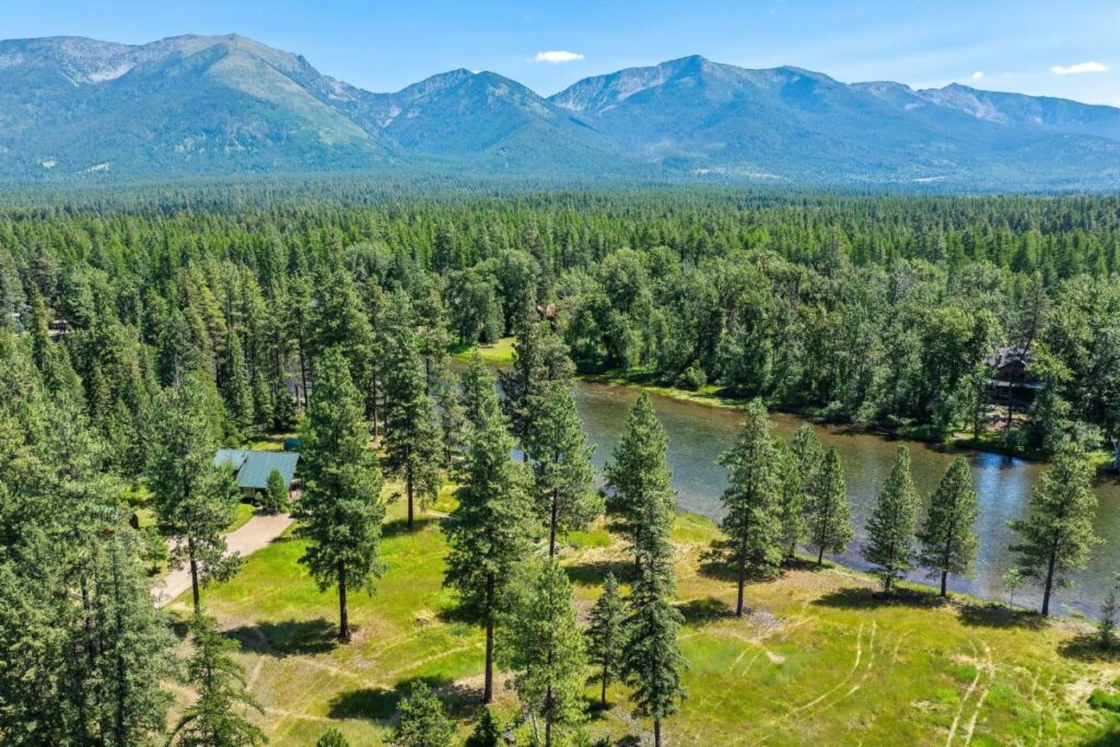 A scenic aerial view of a forested landscape with tall pine trees, a small river or pond, a cabin with a green roof, and mountains in the background under a clear blue sky—perfect for July Journeys and unforgettable Lake Days.