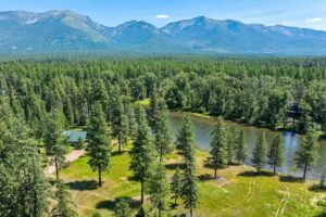 A scenic aerial view of a forested landscape with tall pine trees, a small river or pond, a cabin with a green roof, and mountains in the background under a clear blue sky—perfect for July Journeys and unforgettable Lake Days.