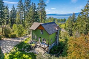 A green two-story house with a wooden deck is surrounded by tall pine trees, evoking winter charm. A lake and mountains are visible in the background under a clear blue sky, perfect for gratitude during the holiday season.