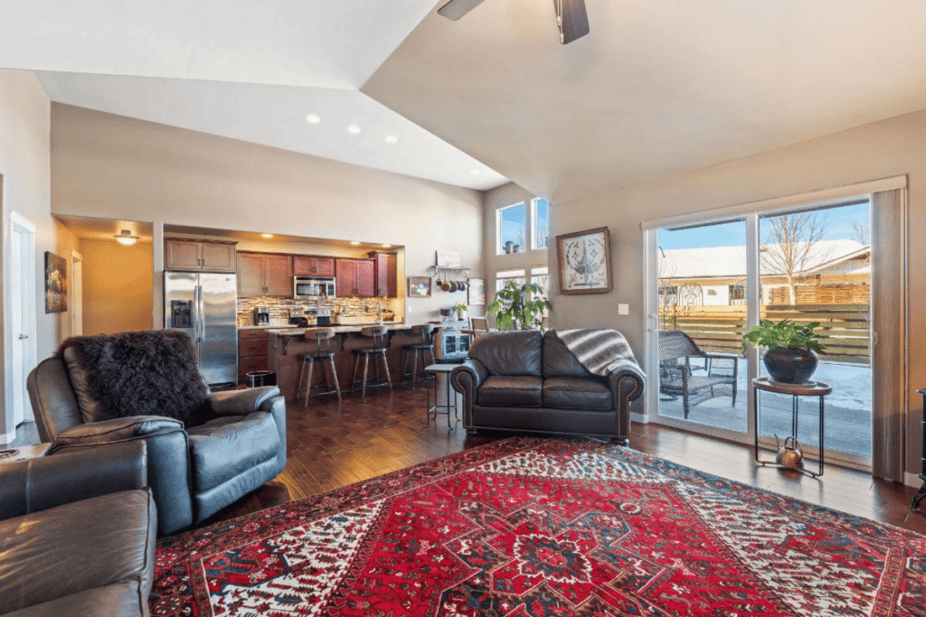 A cozy living room with dark leather furniture, a large red patterned rug, and sliding glass doors leading to a snowy patio. The open kitchen features wooden cabinets and bar stools, with plants by the windows hinting at spring’s arrival.