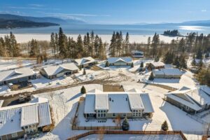 Aerial view of snow-covered houses and trees near a lake, with mountains in the background under a clear blue sky, as love in the air warms this winter scene.