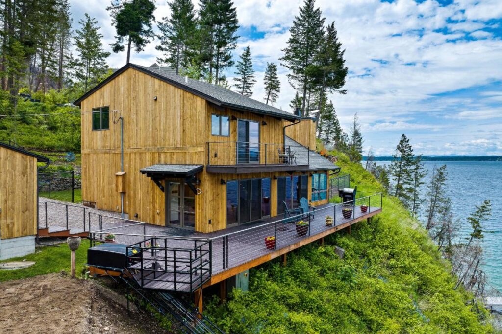 A modern wooden house with large windows and a spacious deck overlooks a scenic lake. Perfect for the HOT Summer Listing Season, this hillside home is surrounded by trees and greenery under a partly cloudy sky.