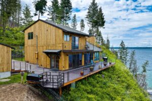 A modern wooden house with large windows and a spacious deck overlooks a scenic lake. Perfect for the HOT Summer Listing Season, this hillside home is surrounded by trees and greenery under a partly cloudy sky.