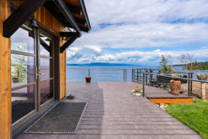 A spacious wooden deck with modern railing overlooks a lake and distant mountains under a partly cloudy sky, attached to a wooden house with large glass doors. Potted plants decorate the deck.
