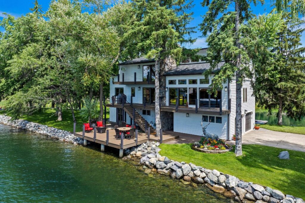 A modern lakeside house with large windows and a stone chimney sits among tall trees, its spacious wooden deck—perfect for Lake Days—outfitted with red chairs and a table, overlooking the clear, calm lake lined with rocks.