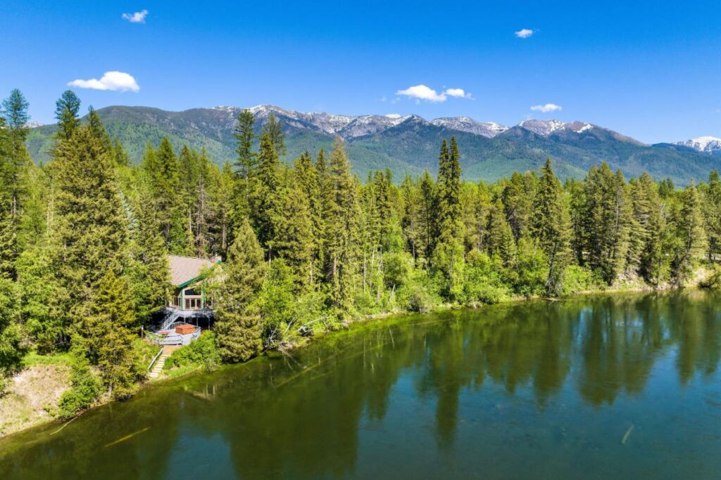 A cabin nestled among tall pine trees sits beside a calm lake, with rolling forested mountains and a clear blue sky in the background.