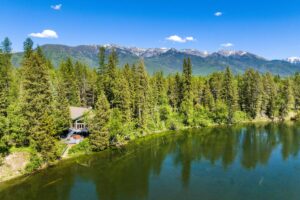 A house nestled among tall pine trees sits beside a calm river, with forested mountains and a clear blue sky in the background, capturing the beauty of fall and the refreshing air of cool days during the changing season.