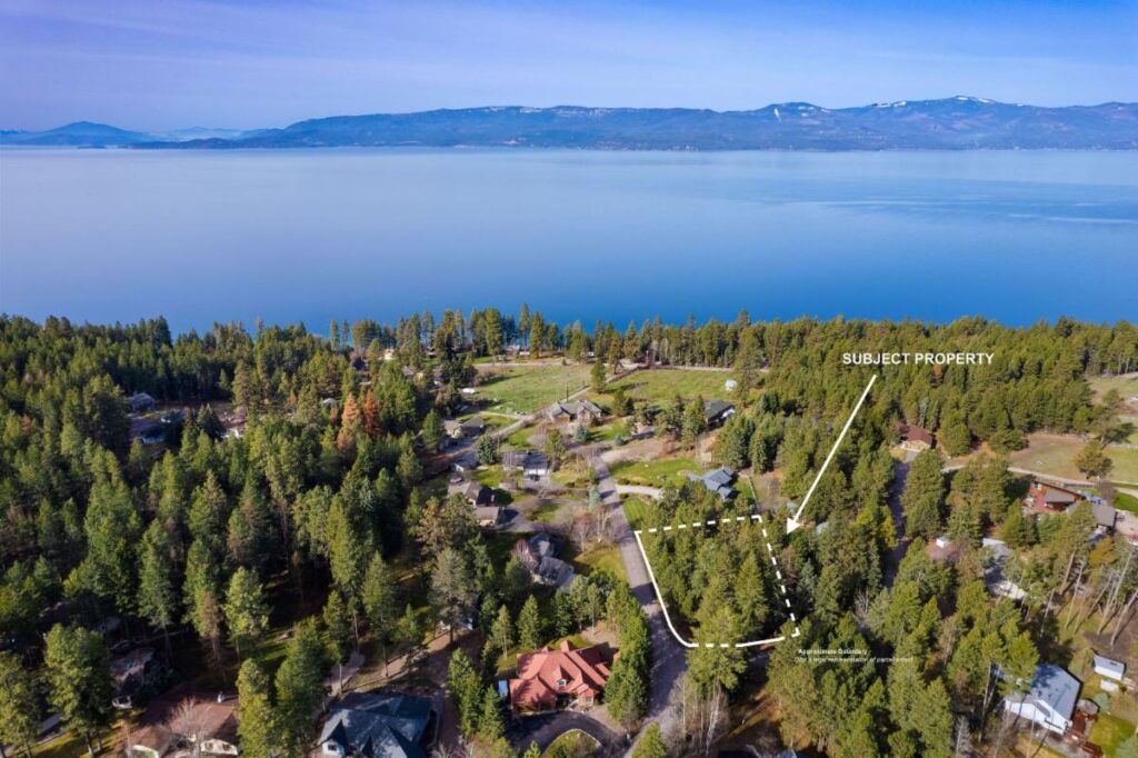Aerial view of a forested lakeside neighborhood with homes among trees; the subject property is outlined and labeled near the shoreline, perfect for the HOT Summer Listing Season, with a large clear lake and distant mountains in the background.