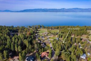Aerial view of a forested lakeside neighborhood in fall with a dotted outline and arrow marking a subject property near the shoreline, surrounded by colorful October trees, houses, and mountains in the background.