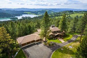 A large house with a brown roof sits among tall evergreen trees on a hillside, overlooking a lake and forested mountains in the distance under a partly cloudy sky. The paved driveway curves in front, hinting at cool crisp days of the changing season.