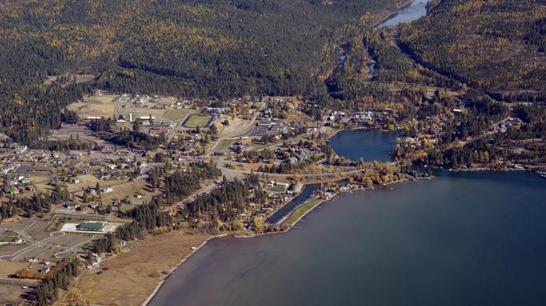Aerial view of a small lakeside town surrounded by forested hills, with scattered houses, roads, and patches of open land near the shoreline in the fresh colors of spring season.