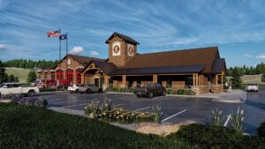 A modern fire station with a stone and wood exterior, a tall clock tower, and red garage doors. American and blue flags fly outside, hinting at June Joys. Cars are parked in the lot and trees surround the building under a blue sky.