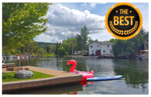 A peaceful lakeside scene with docks, houses, and boats under a partly cloudy sky. A pink flamingo float and paddleboard drift in the water, hinting at changing season and cool crisp days. A The Best award graphic is in the top right corner.